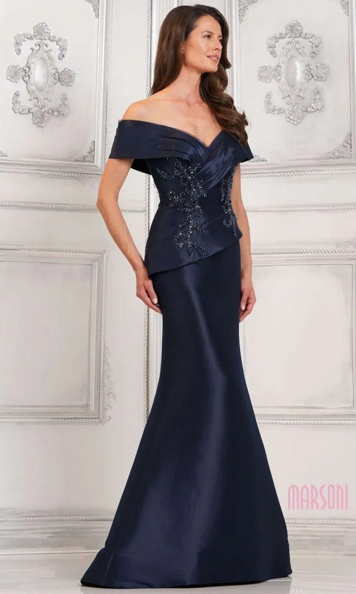 A woman poses against an ornate white background in the Marsoni MV1312 long formal dress, featuring a navy off-shoulder design with beaded embellishments and a fitted mermaid skirt that accentuates her silhouette.